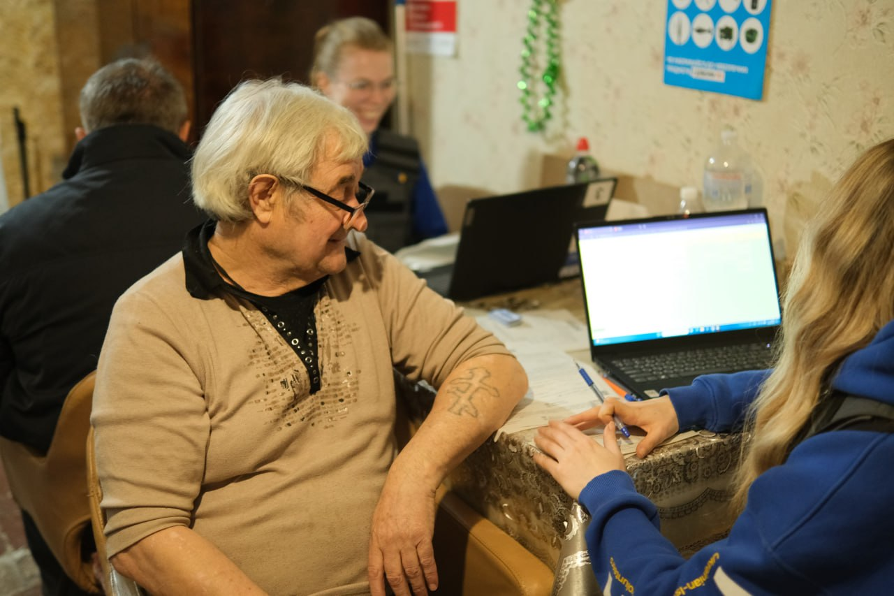 Чоловік похилого віку спілкується з представницею гуманітарної організації, яка вносить дані в ноутбук під час реєстрації або консультації.