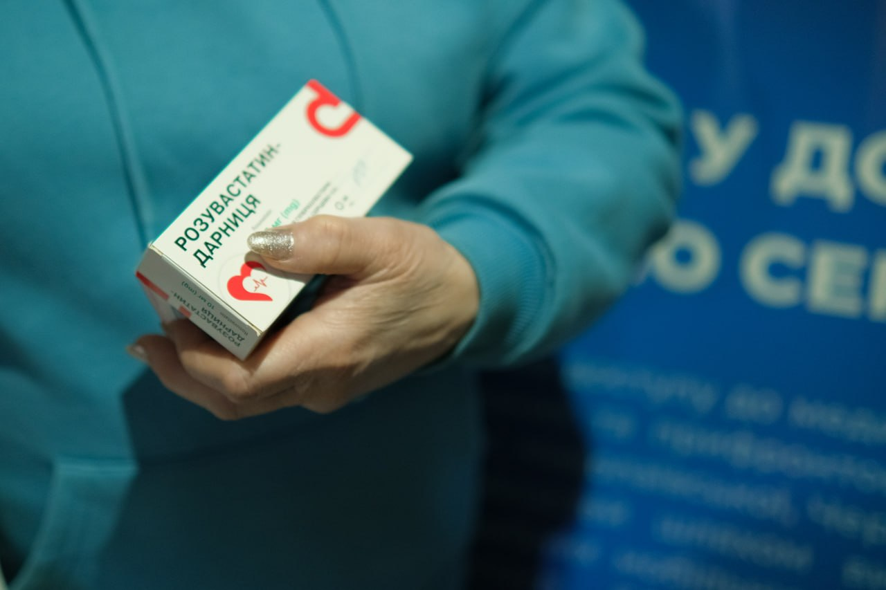 На зображенні жіноча рука тримає упаковку лікарського засобу «Розувастатин-Дарниця» на фоні інформаційного стенда.