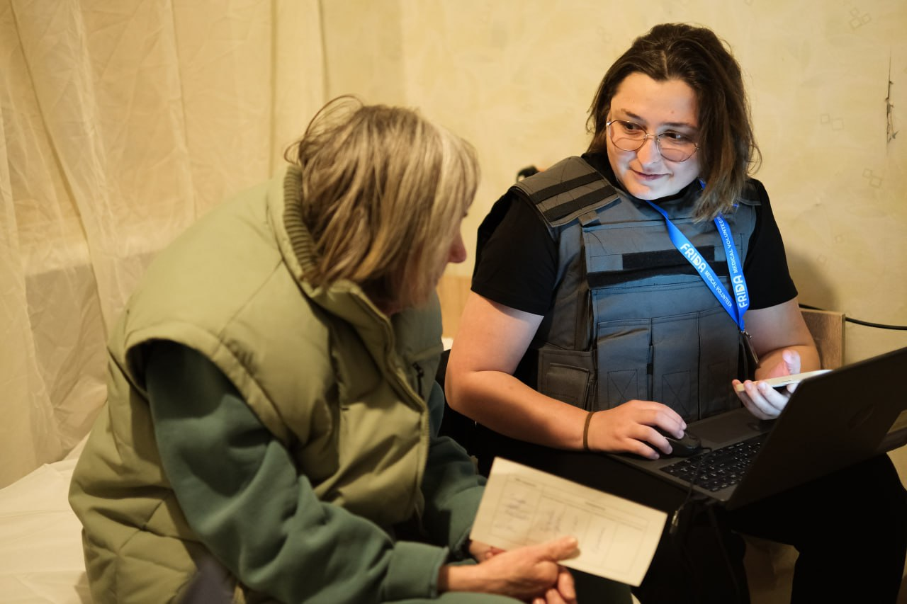 Медична волонтерка в захисному жилеті консультує літню жінку, використовуючи ноутбук, поки пацієнтка тримає в руках медичний бланк.