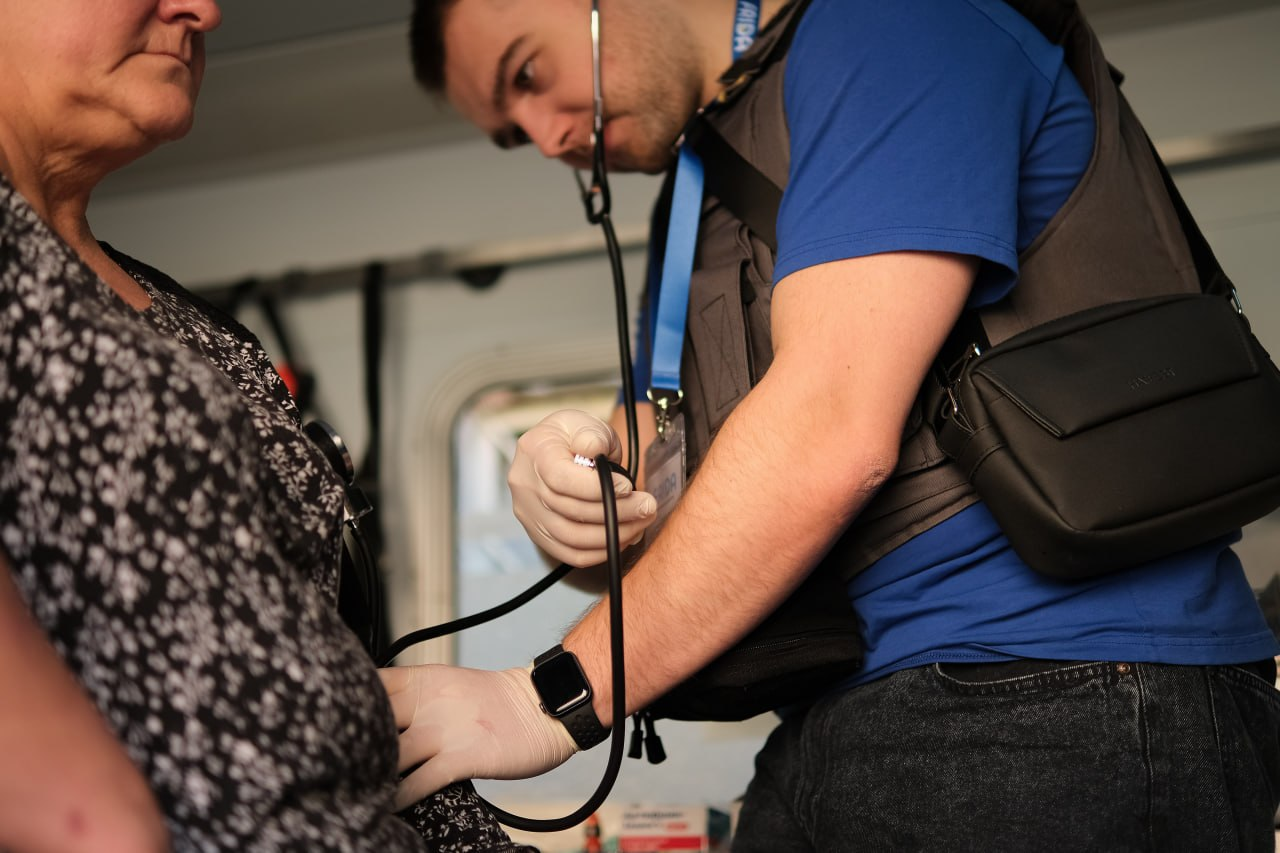 Медичний працівник у захисному жилеті вимірює артеріальний тиск літній жінці за допомогою механічного тонометра та стетоскопа.