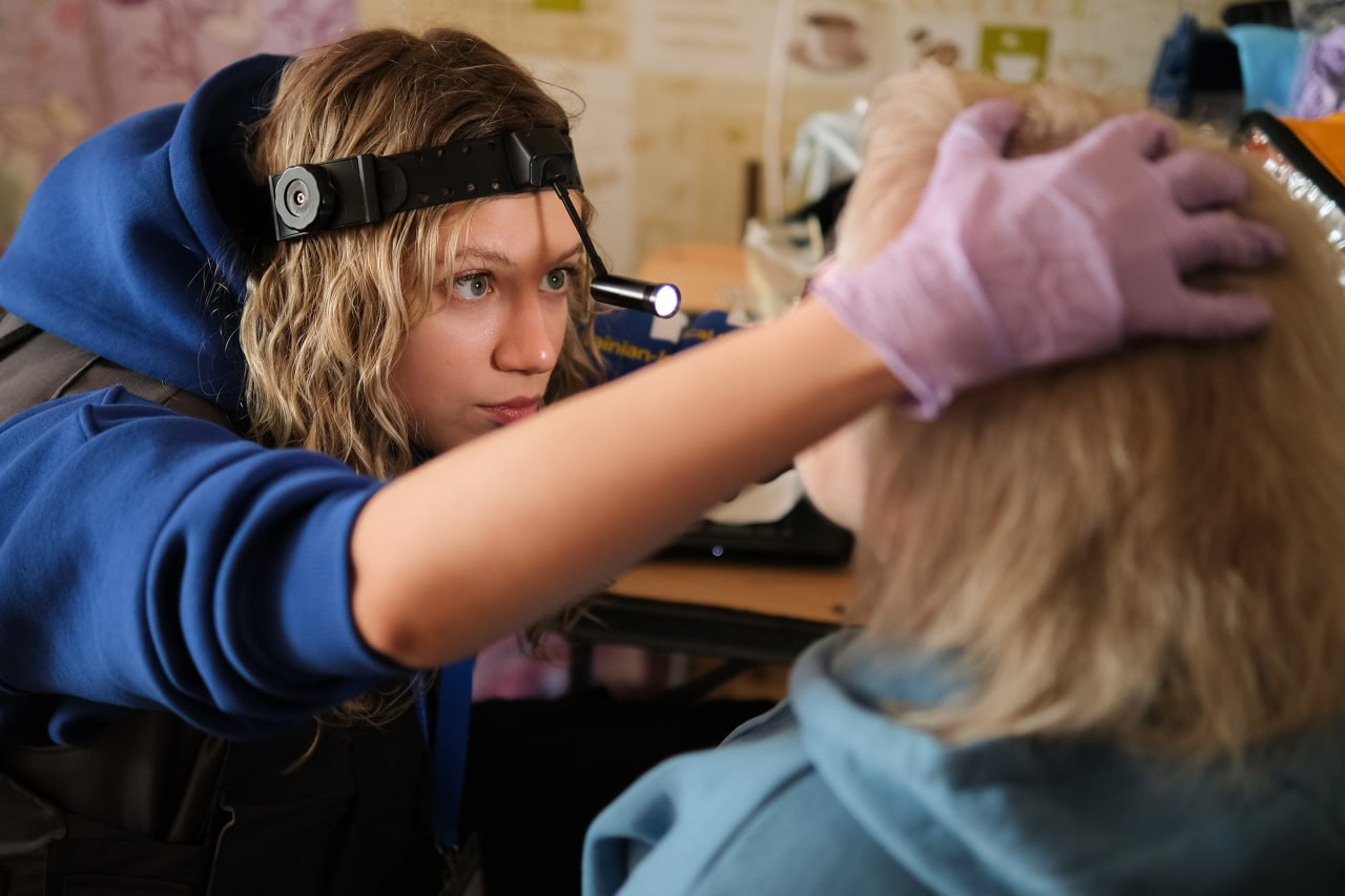 Медична працівниця у захисному жилеті та з налобним ліхтариком проводить огляд голови пацієнтки.