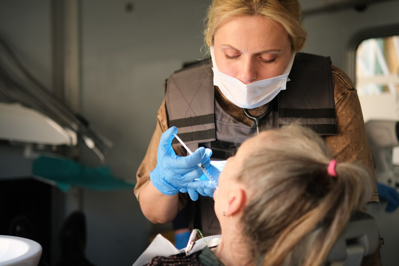 Стоматолог у захисному жилеті та масці робить ін'єкцію пацієнтці в мобільному кабінеті.
