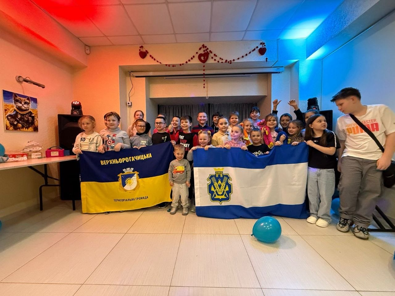 Група дітей позитивно позує для спільного фото в приміщенні, тримаючи прапори Верхньорогачицької територіальної громади та Херсонської області.