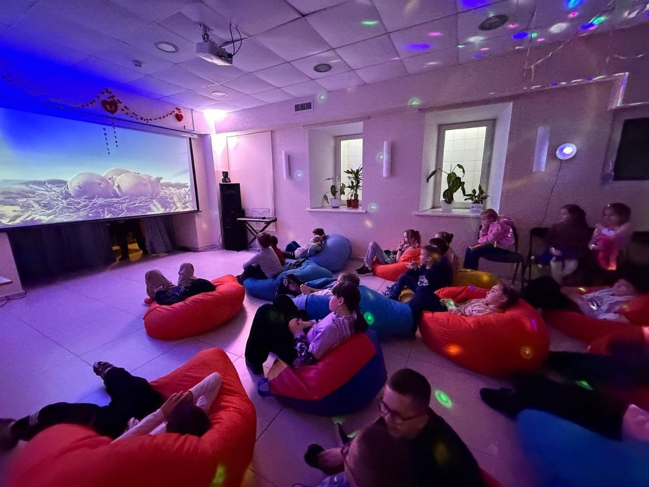 Група дітей відпочиває на різноколірних кріслах-мішках у приміщенні з приглушеним синьо-фіолетовим світлом та дивиться фільм на великому проєкційному екрані.