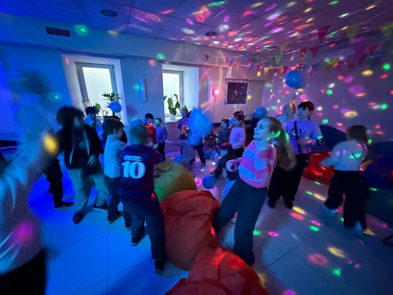 Група дітей активно грається та розважається в приміщенні з яскравим диско-освітленням, кріслами-мішками та святковими прапорцями.