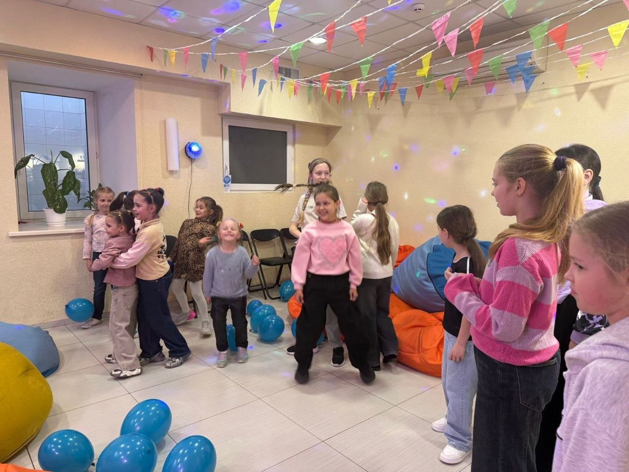 Група дітей активно розважається в ігровій кімнаті, прикрашеній прапорцями та синіми кульками, під світловим супроводом диско-кулі.