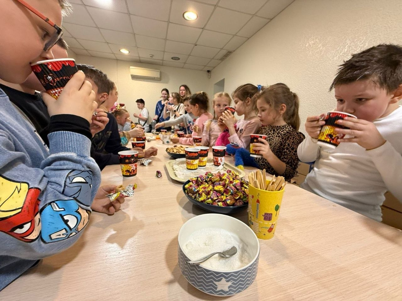 Група дітей сидить за довгим столом у приміщенні, п’ючи напої з паперових стаканчиків та пригощаючись солодощами під час чаювання.