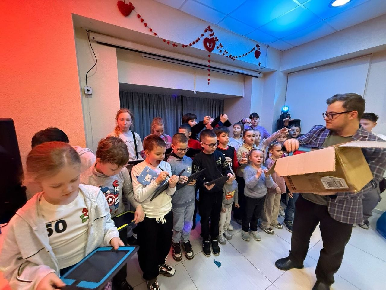 Група дітей захоплено розпаковує подарунки або техніку з великої картонної коробки разом із дорослим помічником у приміщенні, прикрашеному серцями.