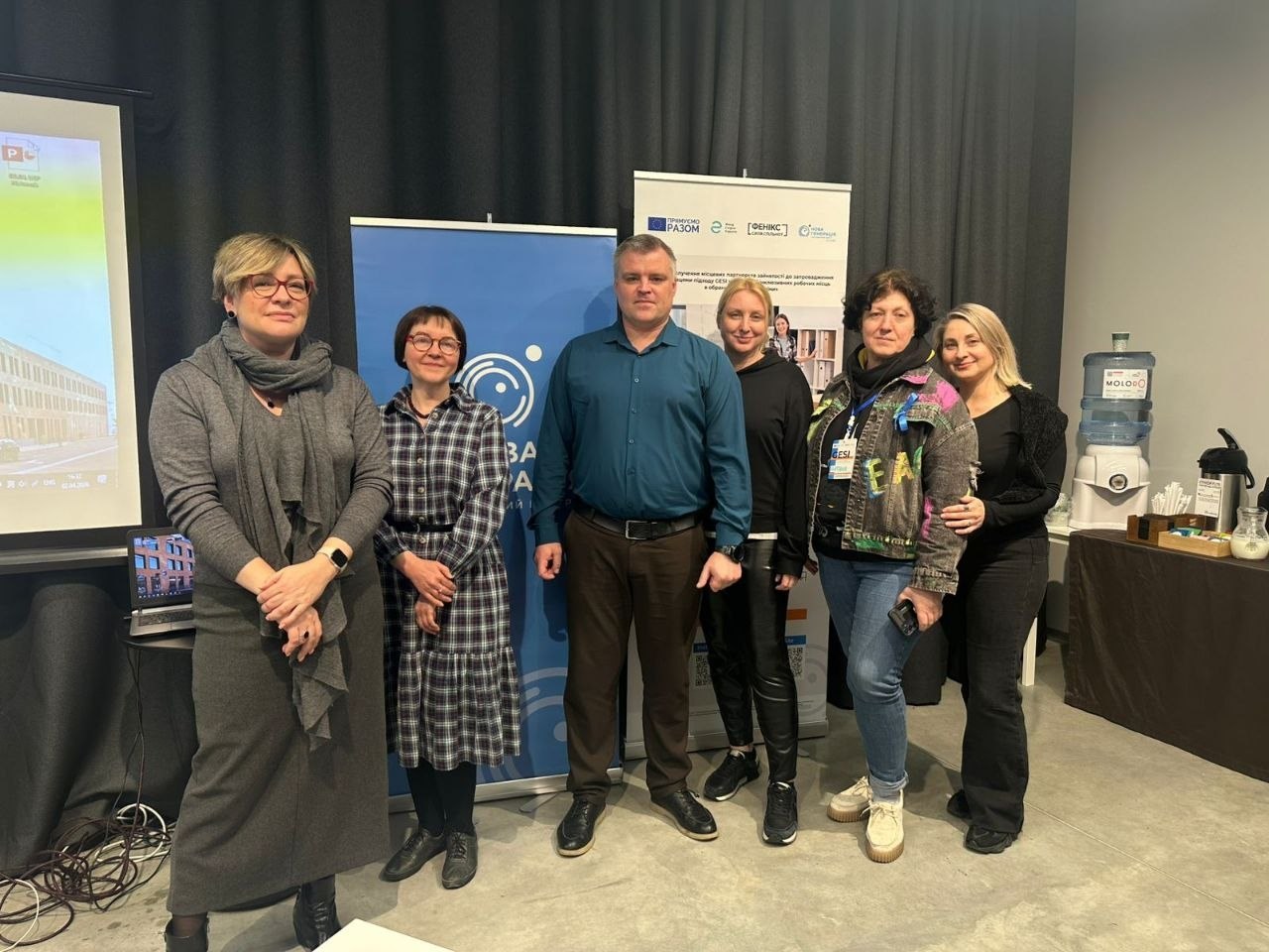 Група людей позує для спільного фото в офісному приміщенні на фоні інформаційних стендів та екрана проєктора.