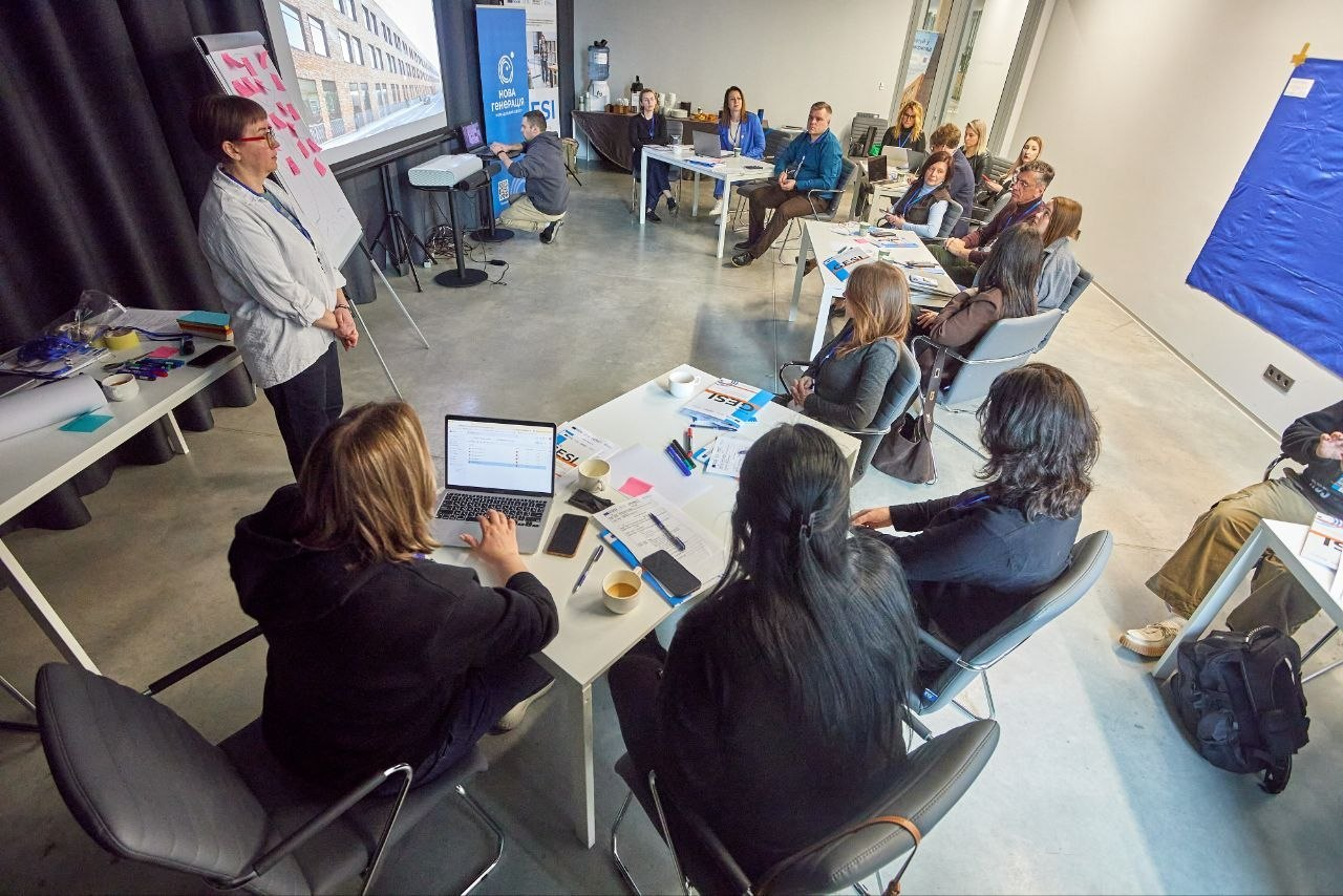 Група людей на тренінгу або робочій зустрічі в сучасному офісі, де модераторка проводить презентацію біля екрана та фліпчарта.