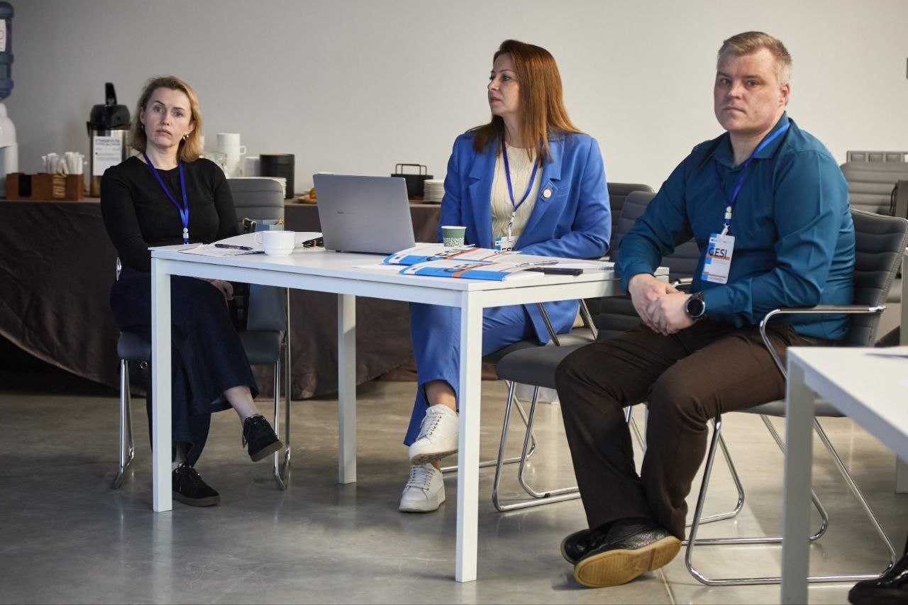 Троє людей з бейджами сидять за білим столом під час офісної зустрічі або навчання в приміщенні.