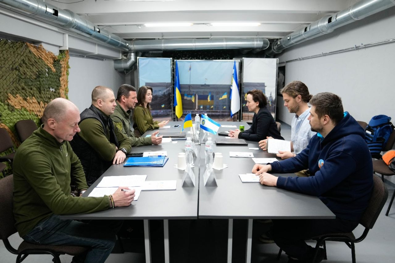 Група людей сидить за довгим столом під час ділової зустрічі або переговорів у робочому приміщенні. Учасники зосереджені, дехто робить нотатки або переглядає документи. На столі та на фоні розміщені невеликі прапори України та міжнародних організацій.