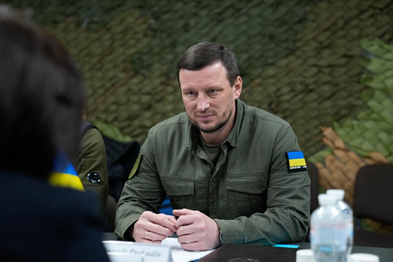 Чоловік у сорочці кольору хакі з нашивкою у формі прапора України на рукаві сидить за столом. Він склав руки перед собою та уважно слухає співрозмовника, який знаходиться поза фокусом на передньому плані. Тло завішене маскувальною сіткою.