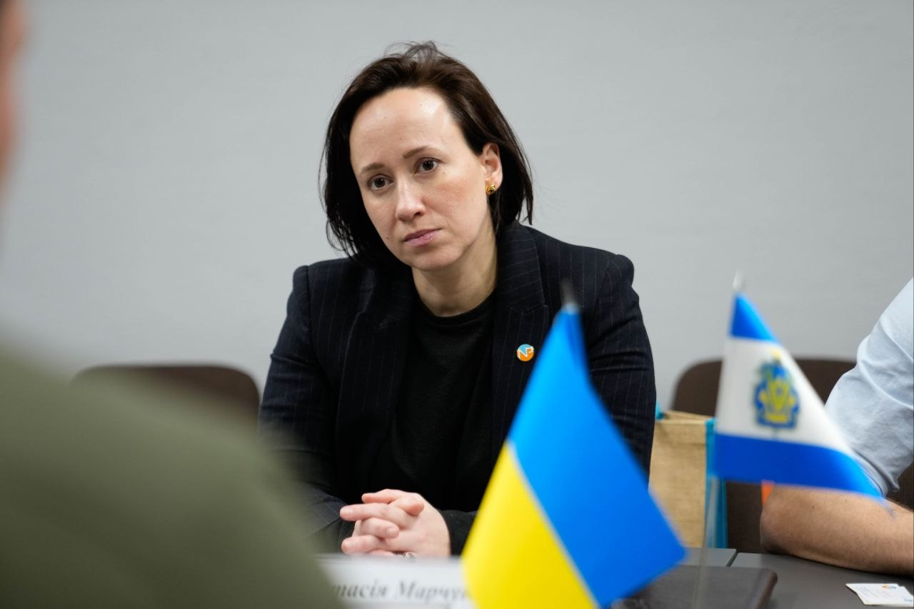 Жінка в темному піджаку зосереджено слухає, сидчи за столом поруч із маленькими прапорами України та міжнародної організації.