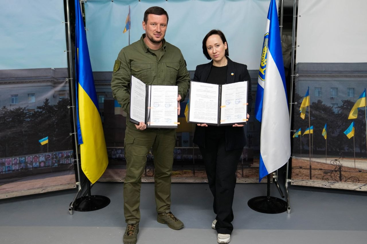 Чоловік у формі кольору хакі та жінка в чорному костюмі стоять поруч, тримаючи відкриті папки з підписаними документами. З обох боків від них встановлені великі прапори — України та міжнародної організації.