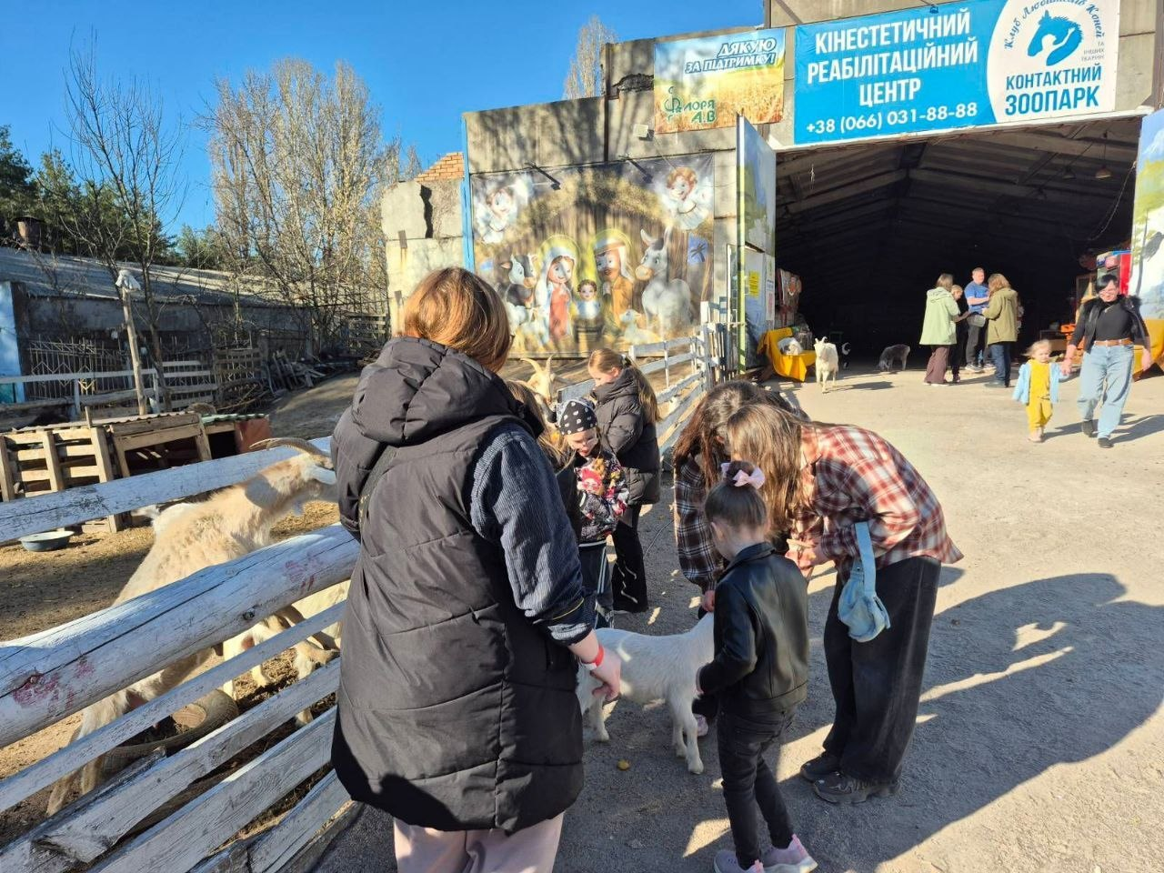 Дорослі та діти взаємодіють із тваринами на території контактного зоопарку та реабілітаційного центру. На фоні видно вольєри, будівлі з інформаційними табличками та відвідувачів на відкритому просторі у сонячний день.