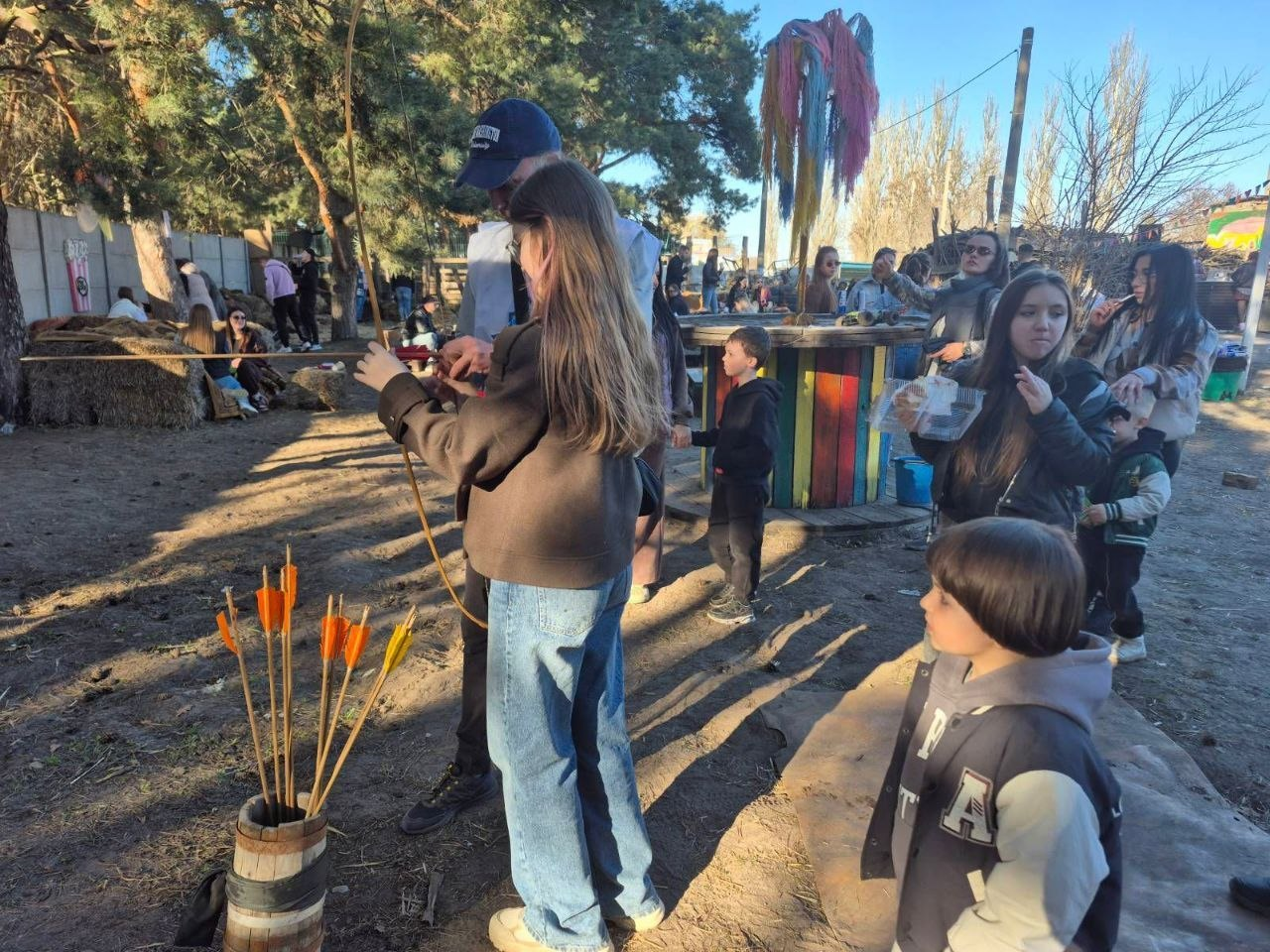 Дорослі та діти проводять час на відкритому майданчику в сонячний день. На передньому плані дитина готується стріляти з лука під наглядом дорослого, поруч стоїть колчан зі стрілами. На фоні видно інших людей, сосни та декоративні елементи.