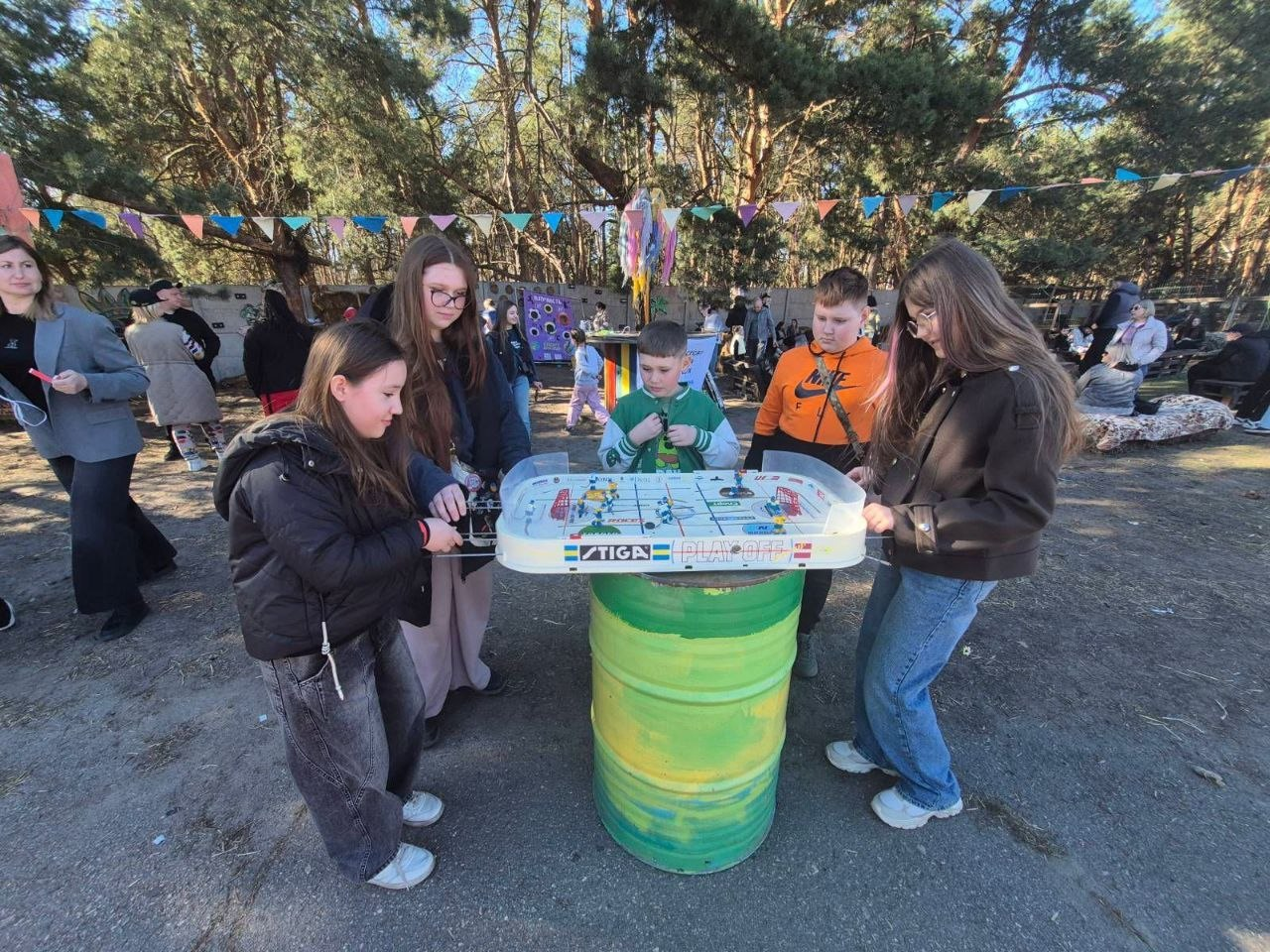 Група підлітків грає в настільний хокей на відкритому повітрі. Ігрове поле встановлене на яскравій зелено-жовтій бочці. На фоні видно сосновий ліс, святкові прапорці та інших людей, які відпочивають у сонячний день.