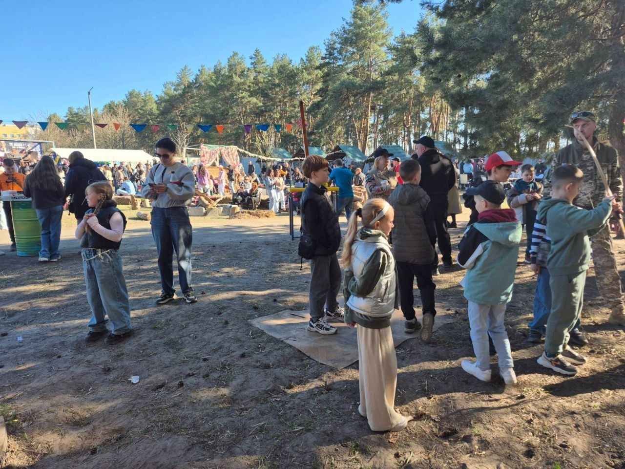 Група дорослих та дітей відпочиває на відкритій локації біля соснового лісу в сонячний день. Люди спілкуються, прогулюються та займаються різними активностями; над майданчиком розвішані кольорові святкові прапорці.