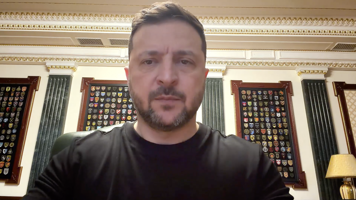 Чоловік у чорному світшоті дивиться прямо в камеру на фоні кабінету з класичним інтер'єром. Позаду нього на стінах розміщені стенди з численними військовими шевронами та емблемами.