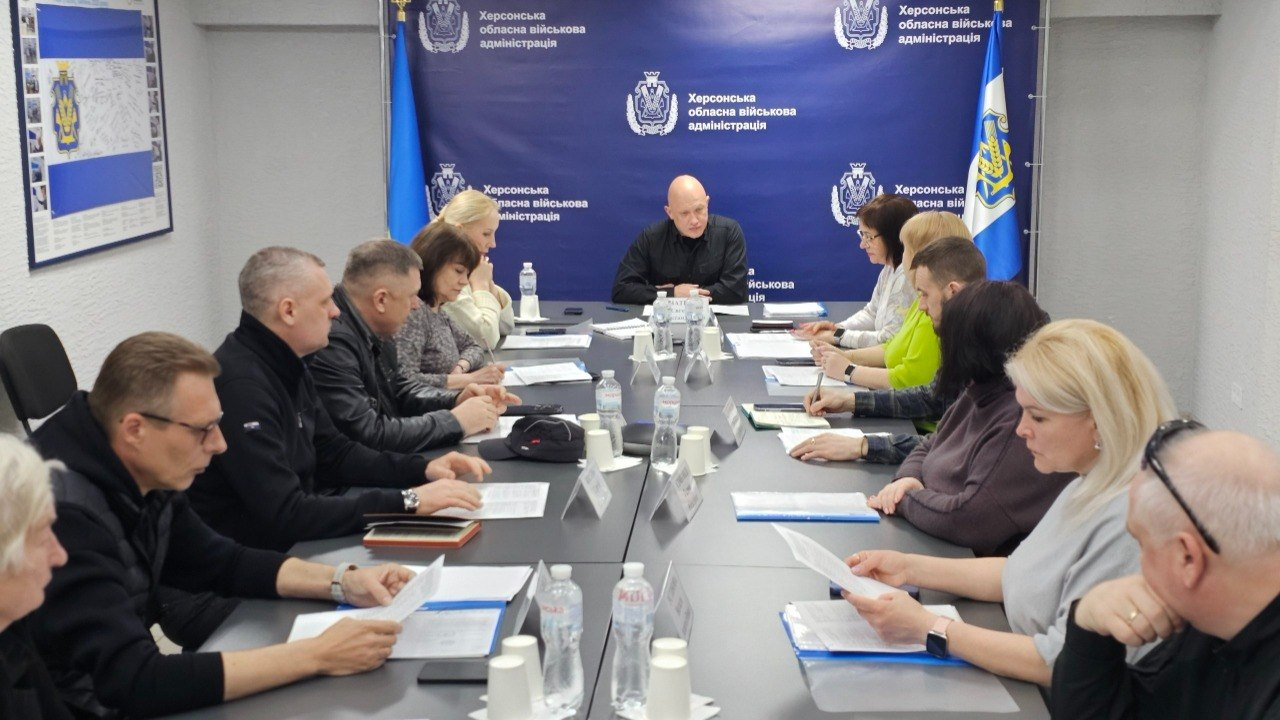 Робоча нарада групи людей за довгим сірим столом в офіційному приміщенні. Учасники вивчають документи на фоні синього брендованого банера та державного прапора України.