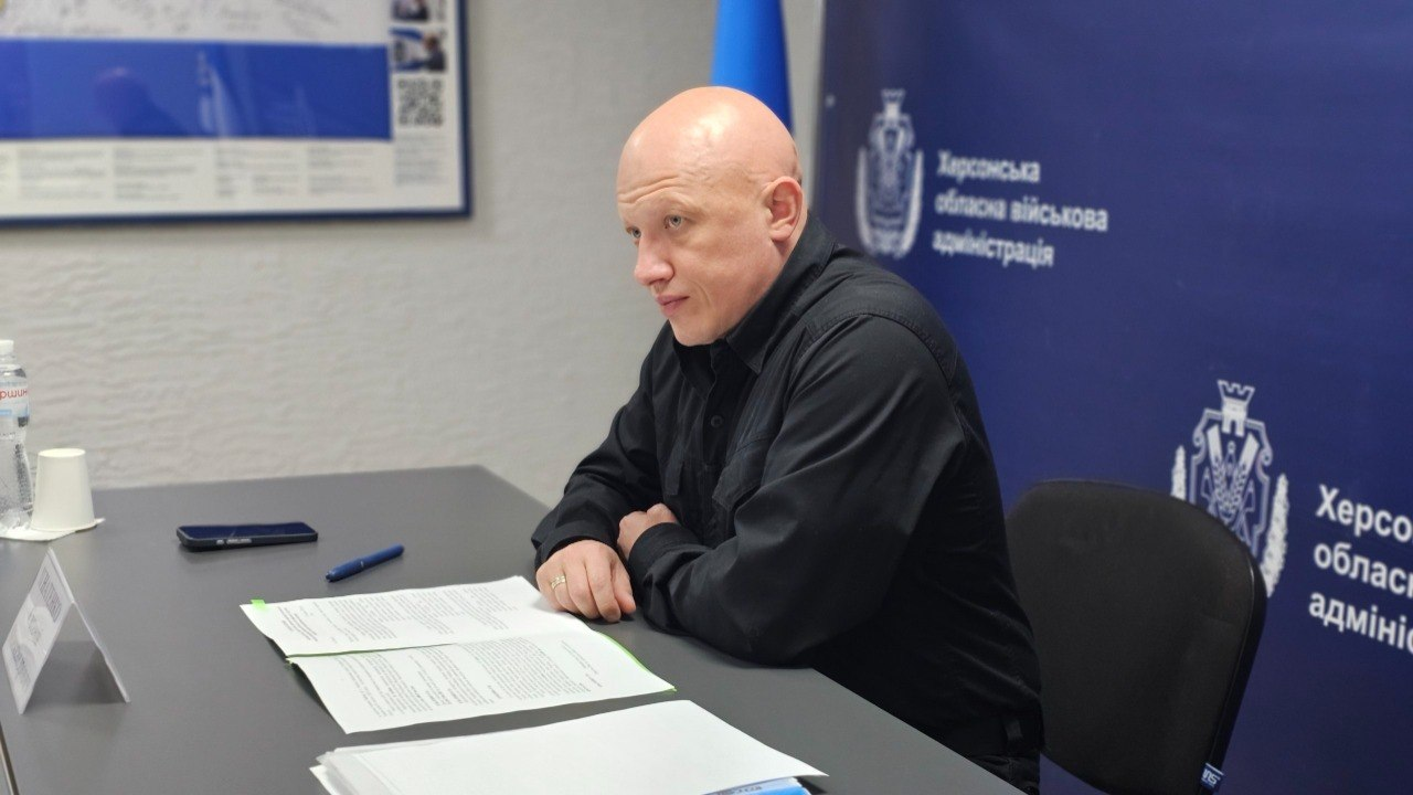 Чоловік у чорній сорочці сидить за робочим столом із розгорнутими документами на фоні синього брендованого банера та державного прапора.