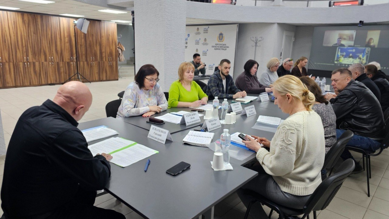 Робоча зустріч групи людей за великим сірим столом в офісному приміщенні. Учасники вивчають документи та працюють із телефонами, а на стіні вдалині видно проекцію відеодзвінка.