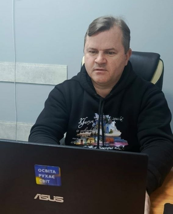 Чоловік у чорному худі працює за ноутбуком в офісному приміщенні. На кришці ноутбука розміщена наклейка з написом «Освіта рухає світ» у кольорах державного прапора України.