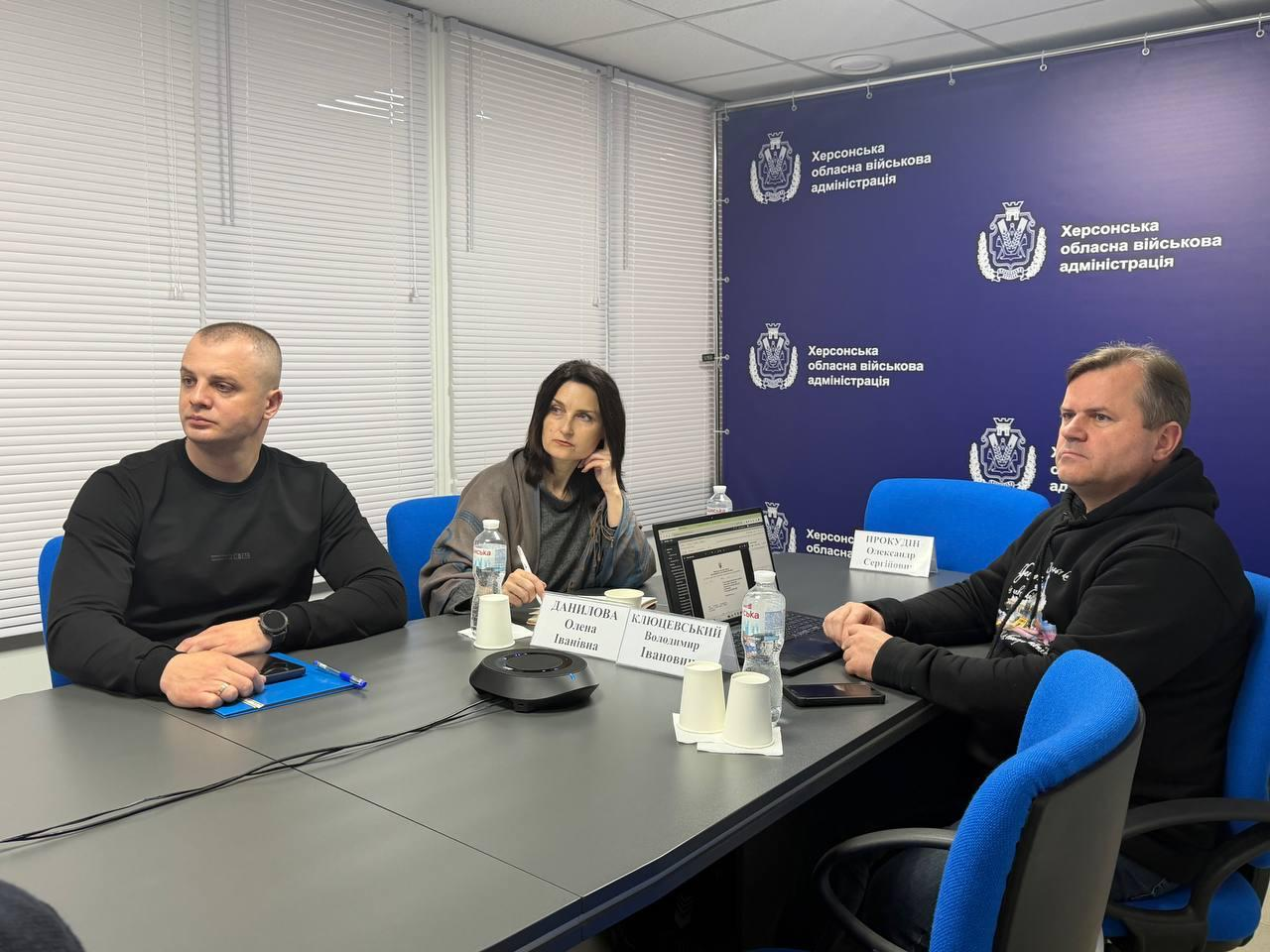 Троє людей у повсякденному одязі сидять за сірим столом під час наради. На столі — мікрофон, таблички та пляшки з водою. Фон складається зі світлих жалюзі та синього банера з офіційною символікою та написами.