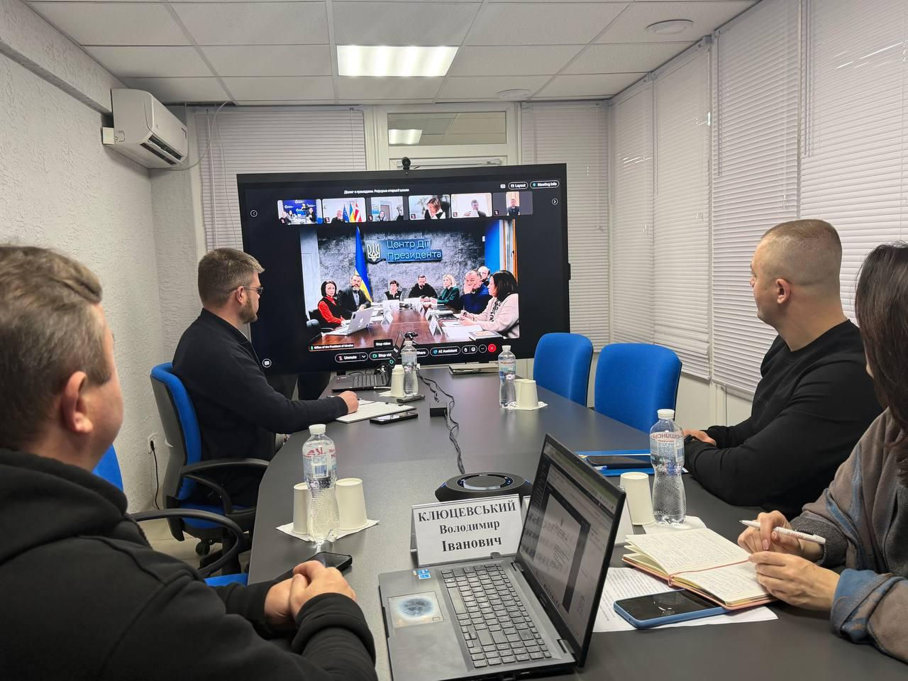 Учасники наради в офісному приміщенні під час відеоконференції з Центром Дій Президента. На великому екрані транслюється онлайн-зустріч, за столом сидять люди з ноутбуком та блокнотами.