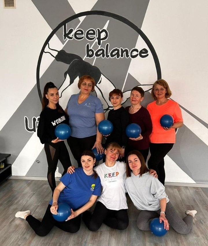 Група з восьми жінок у спортивному одязі позує для спільного фото у фітнес-залі. Деякі з них тримають невеликі сині м’ячі для пілатесу. На фоні — стіна з графічним логотипом та написом «keep balance».