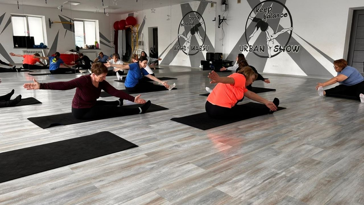 Група людей у спортивному залі виконує вправи на розтяжку, сидячи на килимках. На задньому плані — дзеркальна стіна з великим логотипом «keep balance URBAN SHOW».