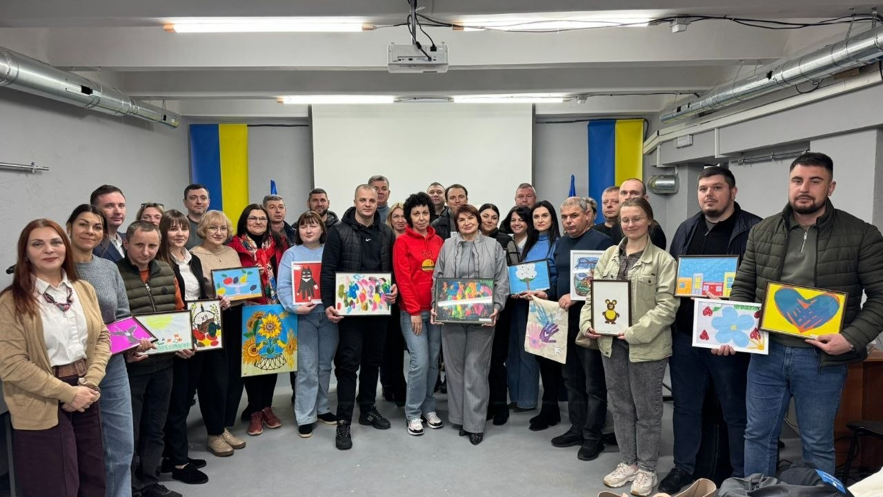 Група людей у приміщенні з українськими прапорами позує для спільного фото, тримаючи в руках яскраві дитячі малюнки в рамках. У залі панує офіційна, але тепла атмосфера творчої зустрічі або благодійного заходу.
