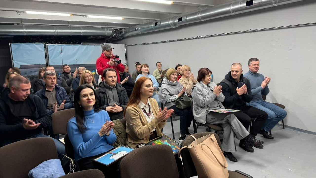 Група людей у цивільному одязі сидить у приміщенні та аплодує. Дехто з присутніх тримає в руках малюнки, на фоні видно технічні комунікації будівлі та банер із зображенням будівлі під державним прапором.