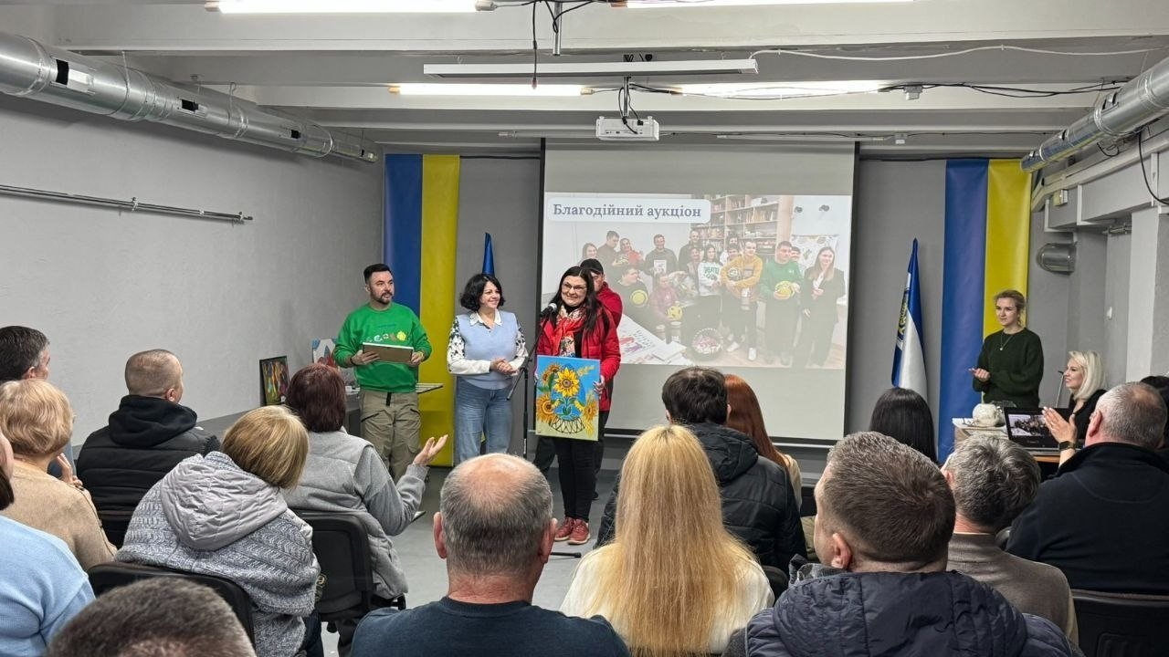 Благодійний захід у приміщенні з українською символікою: жінка перед аудиторією тримає картину з соняшниками. На задньому плані — екран із написом «Благодійний аукціон» та прапори.