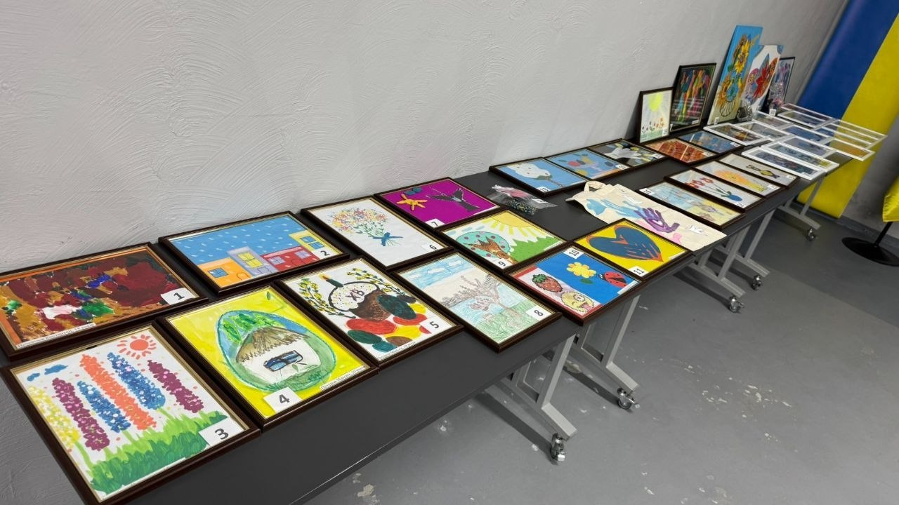 Дитячі малюнки в рамках, розкладені на довгих чорних столах для виставки чи аукціону. Кожна робота має свій порядковий номер. Праворуч на задньому плані видно прапор України.