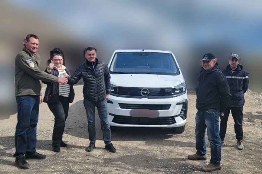 Група людей позує на фоні білого мікроавтобуса Opel. Двоє чоловіків тиснуть один одному руки, а жінка поруч показує великий палець вгору. Подія відбувається на відкритому майданчику з ґрунтовим покриттям.