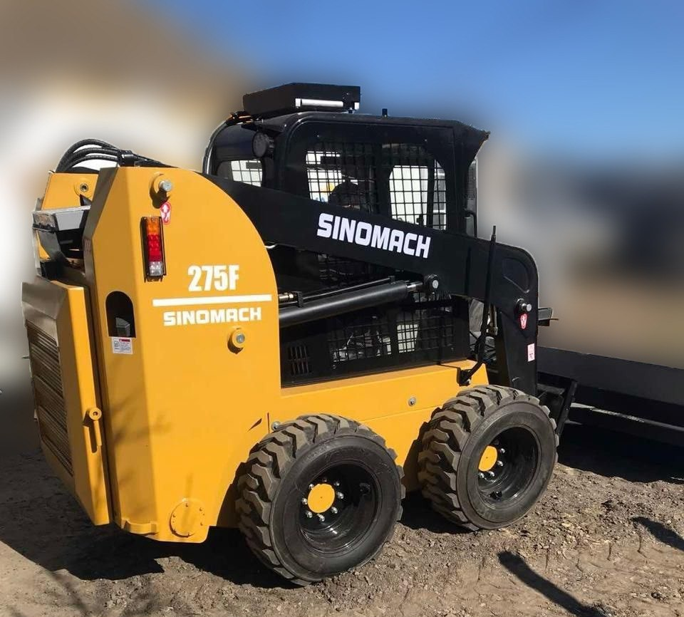 Жовто-чорний мінінавантажувач SINOMACH 275F стоїть на ґрунтовому майданчику в сонячну погоду. Спецтехніка має великі протекторні колеса та закриту кабіну з захисною сіткою.