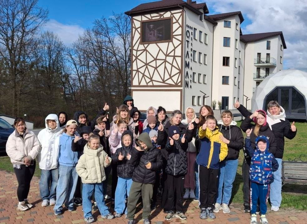 Група дітей та дорослих позує для спільного фото на подвір'ї сучасного готельного комплексу. Учасники посміхаються та показують жест «клас» (великий палець вгору) на фоні багатоповерхової будівлі з геометричним декором.