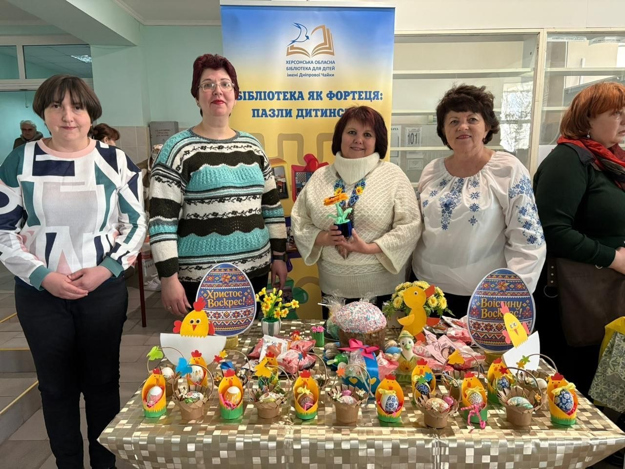 П’ятеро жінок у святковому вбранні, зокрема у вишиванках, стоять за столом із великодніми прикрасами на тлі банера Херсонської обласної бібліотеки для дітей. На столі — святкові кошики з писанками, паперові курчата, декоративні яйця та паска.