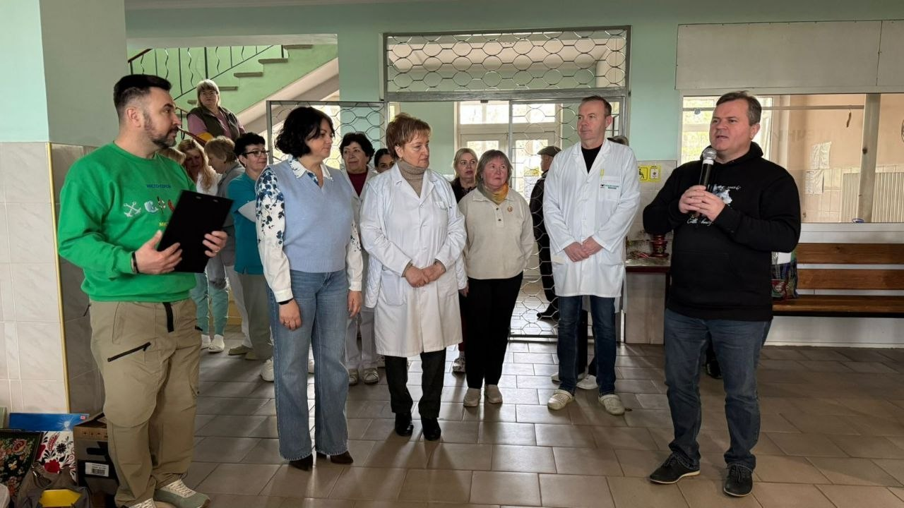 Група людей зібралася у світлому приміщенні, схожому на медичний заклад. Чоловік праворуч говорить у мікрофон, інший ліворуч тримає папку-планшет. Серед присутніх — жінки та чоловіки, деякі з них одягнені в білі медичні халати, інші — у повсякденний одяг.