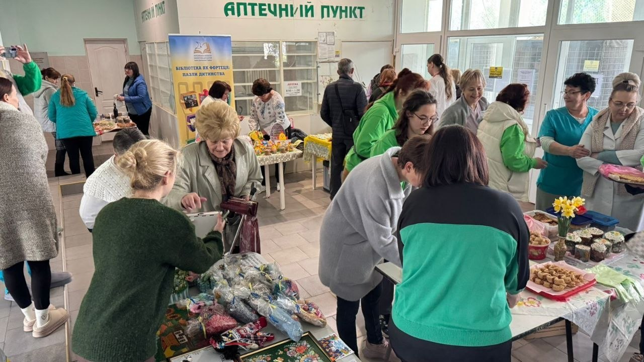Багато людей зібралися у вестибюлі з написом «Аптечний пункт», де розгорнуто благодійний ярмарок. На столах представлені великодні паски, випічка та вироби ручної роботи, а відвідувачі й персонал у медичному одязі оглядають товари.