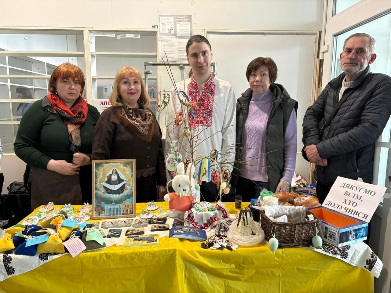 П'ятеро людей стоять за столом, застеленим жовтою тканиною та оздобленим вишитим рушником. На столі представлено великодній декор: паски, писанки, ікону, декоративне дерево та мішечки з сувенірами. Праворуч розміщено табличку з написом «Дякуємо всім, хто долучився».