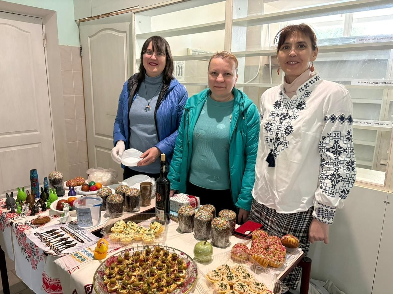 Три жінки стоять за столом із різноманітними частуваннями та великоднім декором. На столі представлені паски, пофарбовані яйця, канапки, тарталетки та випічка, а також фігурки зайців. Одна з жінок одягнена у вишиванку, інші — у повсякденний одяг.