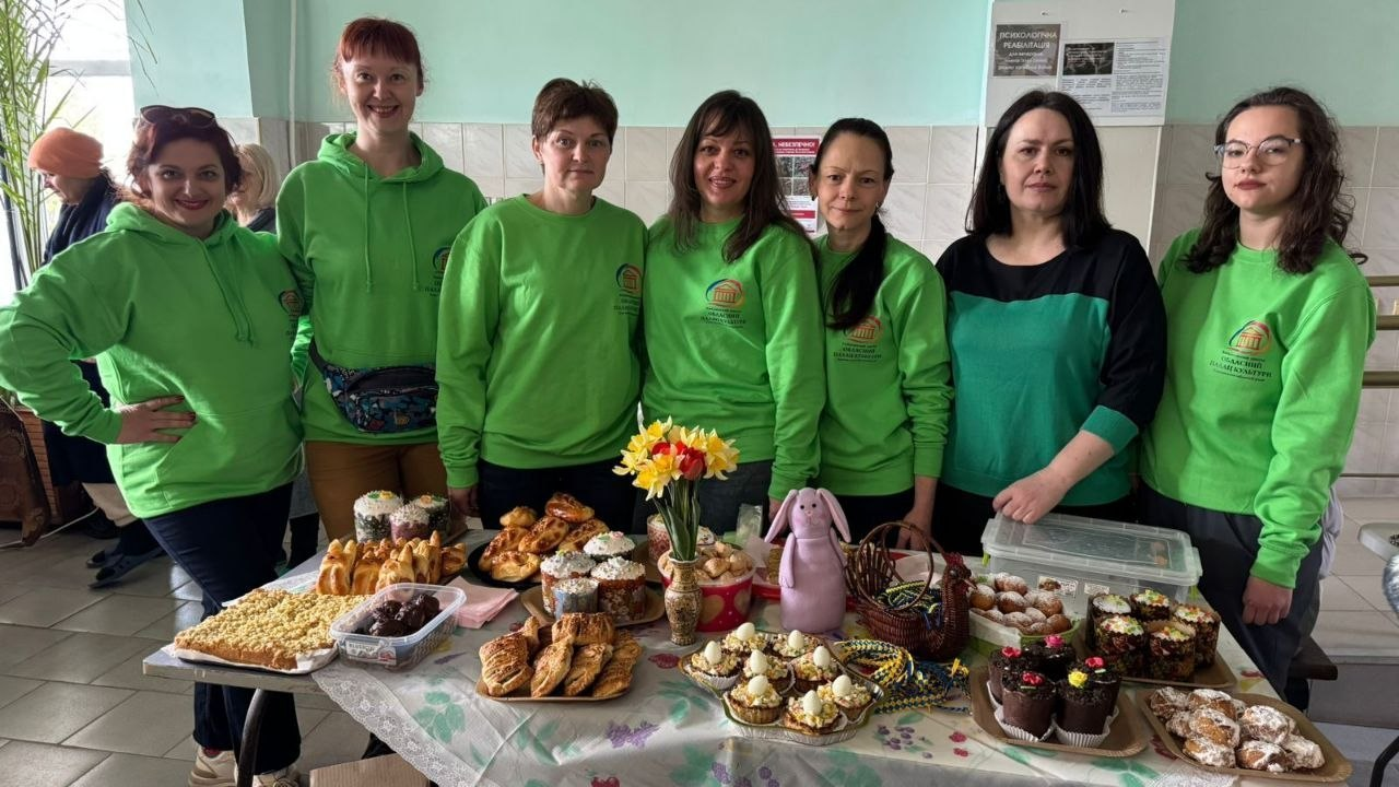 Група з дев'яти осіб у приміщенні з великодніми частуваннями. Сім жінок стоять за святковим столом із випічкою та квітами, більшість у зелених світшотах. На задньому плані ліворуч видно ще двох людей.