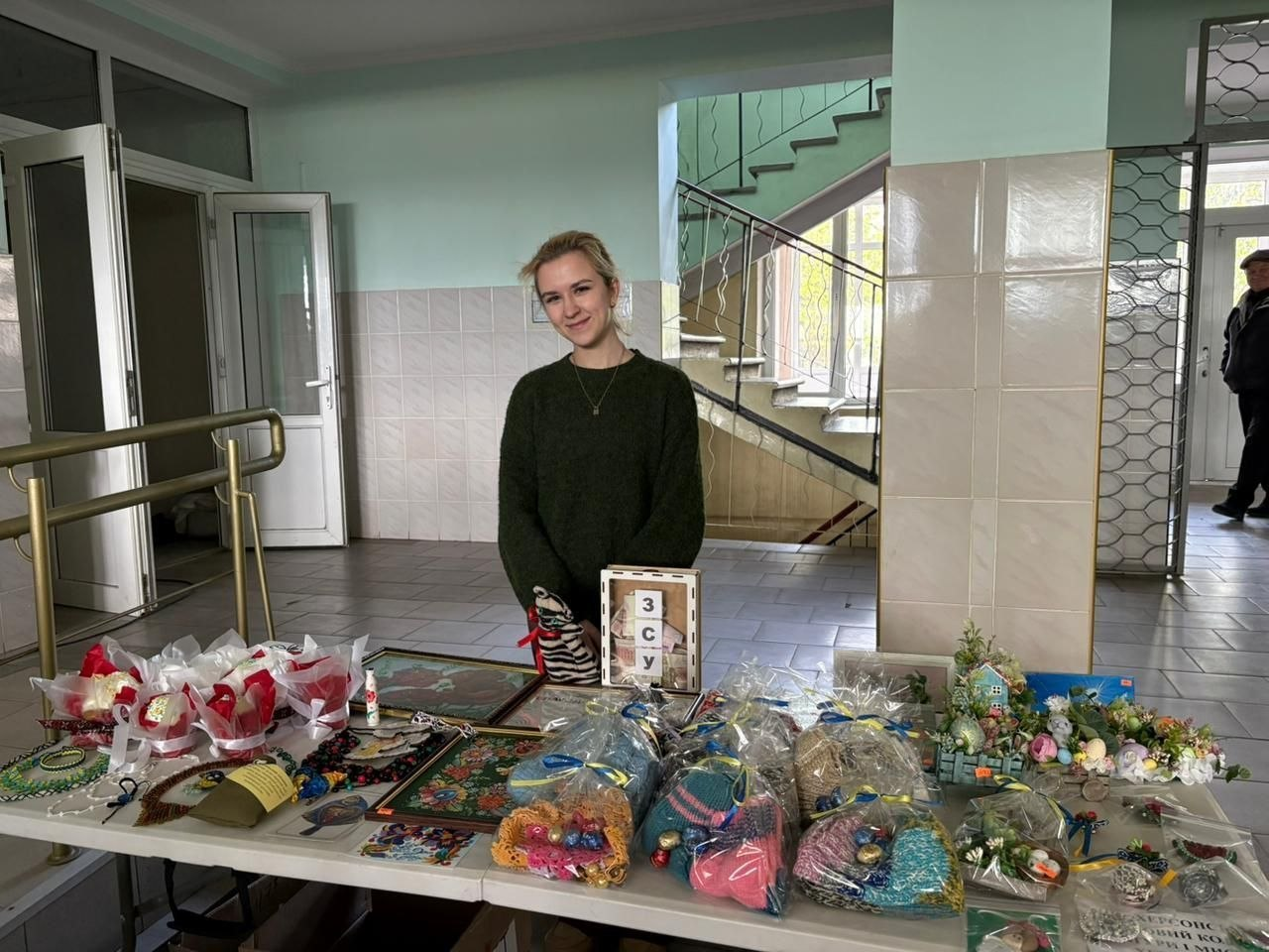 Жінка стоїть за столом із благодійними лотами у світлому приміщенні біля сходів. На столі представлені сувеніри ручної роботи: вишиті картини, в’язані вироби з цукерками, великодній декор та табличка з написом «ЗСУ».