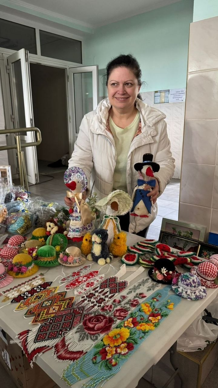 Жінка стоїть за столом, на якому представлені різноманітні вироби ручної роботи: гердани з бісеру, м'які іграшки у формі птахів у капелюхах, декоративні капелюшки-магніти та в'язані вироби. Вона тримає в руках дві авторські ляльки.