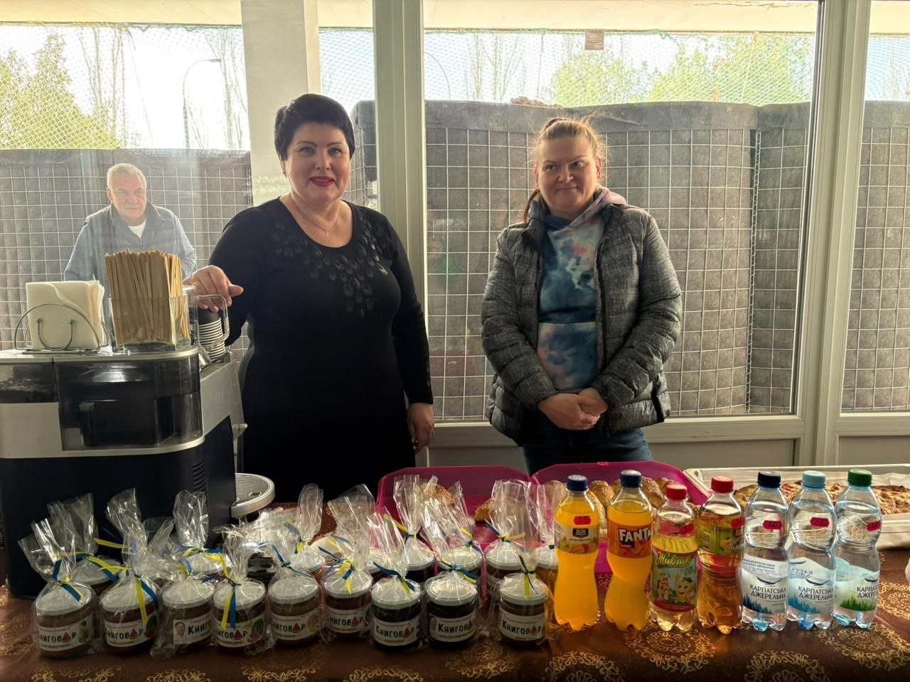 Дві жінки стоять за прилавком із продуктами: баночками, прикрашеними синьо-жовтими стрічками, солодкими напоями та випічкою. Ліворуч на столі розташована кавомашина, а на задньому плані крізь вікно видно чоловіка.