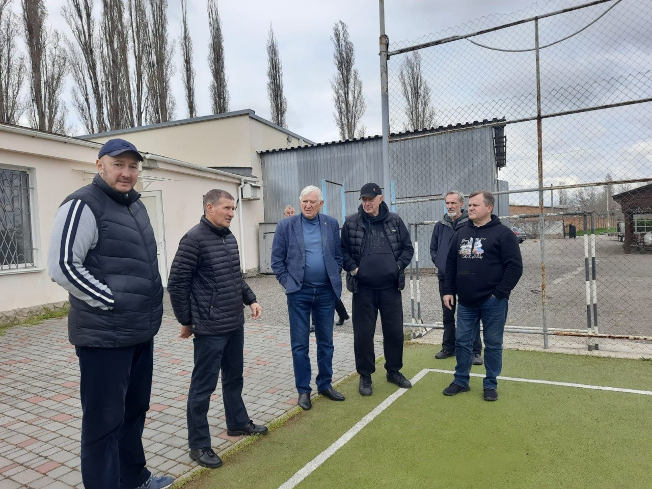 Шестеро чоловіків стоять на спортивному майданчику зі штучним покриттям на фоні огорожі та дерев під хмарним небом.
