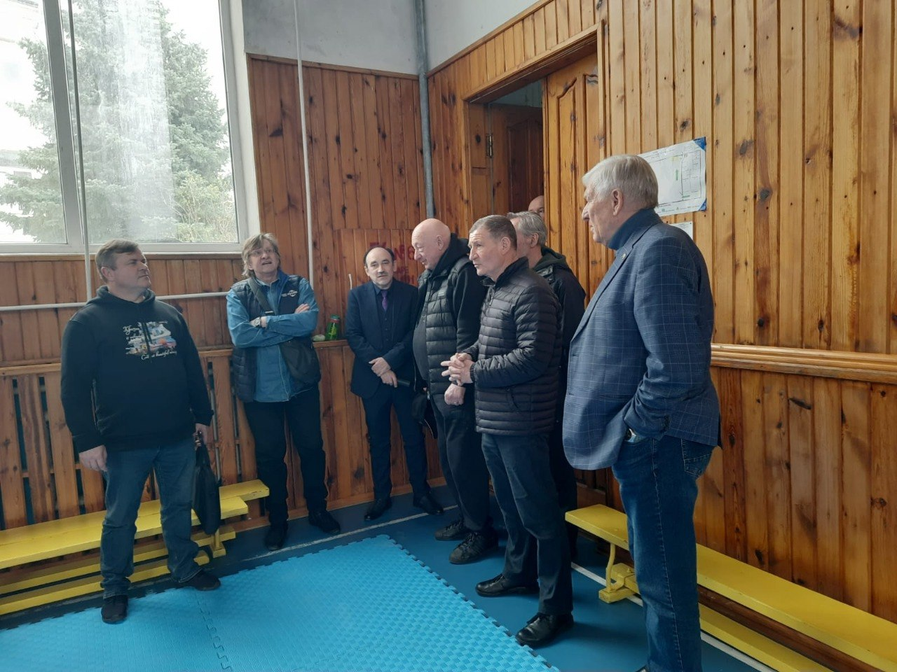 Група з восьми чоловіків спілкується у спортивній залі з дерев'яними стінами, стоячи на синьому маті біля вікна.